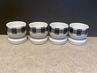 Four white ceramic egg cups with black and white checkered bands arranged in a row on a countertop, viewed from the front.