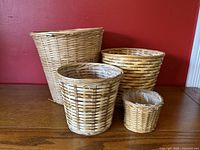 Four wicker baskets displayed on a wooden surface against a red background. Two baskets are larger and two smaller ones, one basket is lined with clear plastic.