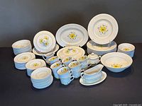 Full set arranged showing all the different dishes, bowls, cups and serving pieces in white with yellow flowers and basketweave texture.