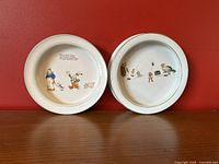 Two white ceramic children's bowls with green trim, each featuring colorful nursery rhyme illustrations inside depicting characters and scenes. One bowl shows a person with a clown and pig, the other multiple children and animals.