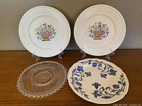Four plates and one glass serving dish displayed on wooden surface and stands. Three white Wedgwood Edme Borrowdale plates have colorful floral baskets centered with ribbed edges. One white Royal Staffordshire Windsong plate features blue floral and scroll design. Clear glass oval dish has etched circular pattern and scalloped edge.