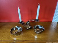 Full view of two brushed steel candle holders each holding a white taper candle, alongside two wavy rim silver glass bowls on wooden table with red background