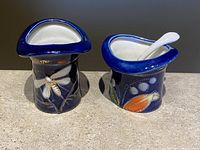 Two cobalt blue Victorian style porcelain kitchenware items on a beige surface: a top hat toothpick holder with dragonfly decoration and an open sugar bowl with a spoon inside.
