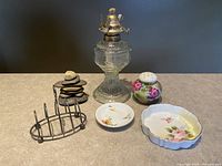 Front view of the mixed collectible lot including glass oil lamp, stone sculpture, ceramic salt shaker, small floral dishes, and metal toast rack on table surface.