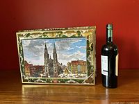 Front view of the large E. Otto Schmidt embossed cookie tin with St. Lorenz church image next to a wine bottle for scale.