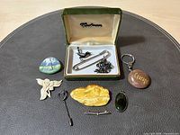Green velvet jewelry box open showing assorted vintage and artisan jewelry items including brooches, pendant, a keychain, and a small pin on a dark surface