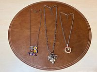 Three silver-tone charm necklaces laid flat on a brown circular surface showing overall length and pendant styles