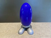 Front view of blue egg-shaped bedside lamp with silver rounded dinosaur-like feet base on a flat surface against a dark background.