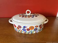 Oval ceramic casserole dish with colorful floral and butterfly pattern with lid, on a wooden surface against a red background.