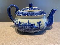 Side view of the ironstone teapot showing decorative blue flow pattern with building and landscape motif and blue handle and spout
