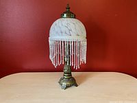 Lamp with frosted glass dome and beaded fringe shade, metal ornate base against red wall.