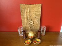 Full view of all items including carved wall plaque, candlesticks, ceramic shaker, coasters, and glasses against red backdrop.