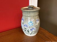 Full view of the pottery jar showing shape, lid, and floral design against red and brown background.