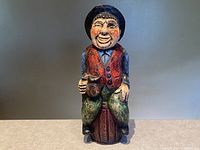Front view of hand carved wooden folk art wine bottle holder shaped as a smiling man sitting on a wine barrel holding a wine mug.