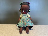 Full view of handmade cloth doll seated against a neutral background showing paisley skirt and dark brown fabric body.