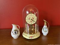 Kern mantle clock under glass cloche with two small German souvenir pitchers beside it on wooden surface against red wall.