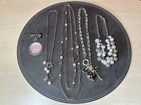 Photo displaying the four fashion necklaces arranged on a black round placemat. Items include a multi-strand faux pearl necklace, a silver-tone chain with black heart pendant and charms, a delicate long chain with small pearls and geometric beads, and a silver link chain with a pink medallion pendant.
