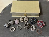 All 11 fashion rings arranged in front of a gold-tone presentation box on a dark surface, showing variety in size, color, and style.