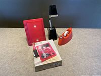 View of three items on a surface: pink picture frame, black adjustable lamp, orange tape dispenser