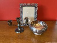 Set of metal items on a wooden surface against a red wall: an ornate photo frame, Marlboro bowl, and 3-arm candlestick with felt bottom
