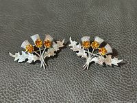 Pair of sterling silver Scottish thistle brooches with three citrine-colored glass stones each, shown side by side on a textured black surface.