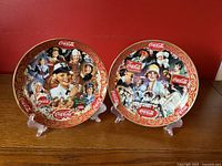 Two 8-inch Coca-Cola collector plates displayed upright side-by-side on clear stands, showing detailed retro illustrations and Coca-Cola logos within ornate red and gold borders.