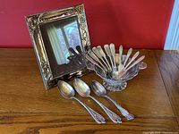 Overall view of silver picture frame, silver overlay glass decorative bowl with handles, and assortment of spoons and spreaders laid on a wooden table.