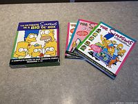 Photo showing the main box and three accompanying books featuring The Simpsons characters on the covers laid out on a table.