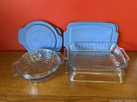 Three vintage Anchor Hocking glass baking dishes with lids shown, including one round dish with glass lid and two rectangular dishes with blue plastic lids on wooden surface against red wall.