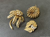 Three gold tone vintage brooches on black leather background showing detailed textures, faux pearls, and rhinestones.
