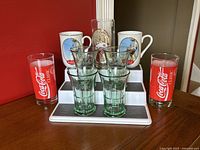 Full set of Coca-Cola drinkware with two Norman Rockwell ceramic mugs, four green Libbey glasses, and two red Coca-Cola tall glasses against a wood table and red wall.