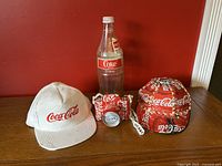 Group photo of three items: white corduroy Coca-Cola snapback hat, handmade camera made from Coca-Cola cans, and vintage 1.5L glass Coca-Cola bottle, set against a red wall.