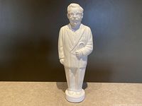 Full front view of the white plastic Colonel Sanders figure bank showing him dressed in a suit holding a cane, standing upright on a circular base.