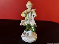Close-up of one male porcelain figurine playing flute in Victorian style attire, seated on a white ornate base, hand-painted details visible.