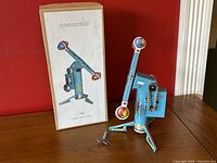 Photo of the blue tin wind-up rocket ride toy next to its original box and winding key, standing on a wooden surface with a red wall background.