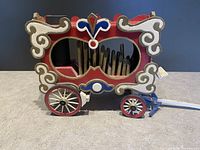 Front and side views of the vintage wooden circus calliope showing decorative scrollwork, metal pipes inside, wooden wheels, and pull handle.