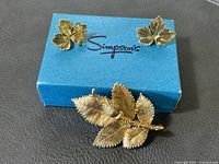 Close shot of gold-tone textured rosebud and leaf brooch next to a pair of gold-tone maple leaf clip-on earrings on a blue Simpson's box.