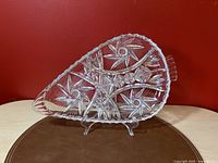 Oblique angle side view of the clear crystal divided serving dish showing intricate starburst patterns and flared handle, placed against a red background on a light wood surface with a brown pad beneath.