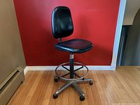 Full view of the black vinyl drafting stool with high backrest, swivel seat, metal foot ring, and metal caster base against a red wall.