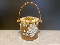 Front angle view of the vintage ceramic biscuit jar showing the golden glaze and white raised grape design with woven handle.