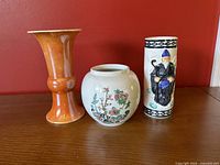Three vintage porcelain vases displayed against a red background including an orange Grimwades vase, a white floral Sadler vase, and a Cylindrical vase featuring an old wise man figure Made in Japan.