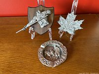 Front and angled view of three vintage metal items: javelin thrower plaque on wood, Canadian maple leaf metal plaque, and round metal ashtray from Portugal.