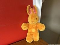 Front view of the vintage mohair bunny rabbit plush showing yellow-orange fur with pink crushed velvet inner ears, standing on a wooden surface against a red wall.