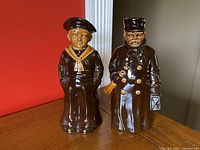 Full view of two ceramic decanters shaped as navy figures placed on wood surface against red and white background.
