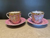 Front view of two pink porcelain tea cups with gold floral and leaf pattern paired with matching pink saucers, scalloped edges and gold detailing