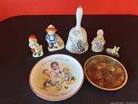 All porcelain items including commemorative plate, bell, figurines and decorative bowl shown together on black surface before red background.