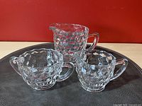 Three pressed glass items arranged on a round dark surface against a red background: a sugar bowl, a creamer, and a small pitcher, all with a diamond cube raised pattern.