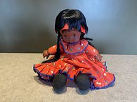 Front view of the seated vintage children's doll with orange dress, black braided hair, and freckles.