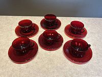 Six dark ruby red glass teacups each paired on saucers shown with the cups placed on top of the saucers on a countertop.