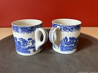 Two teacups side-by-side with handles facing each other, showing detailed blue willow pattern and ornate handles with key-fret design.